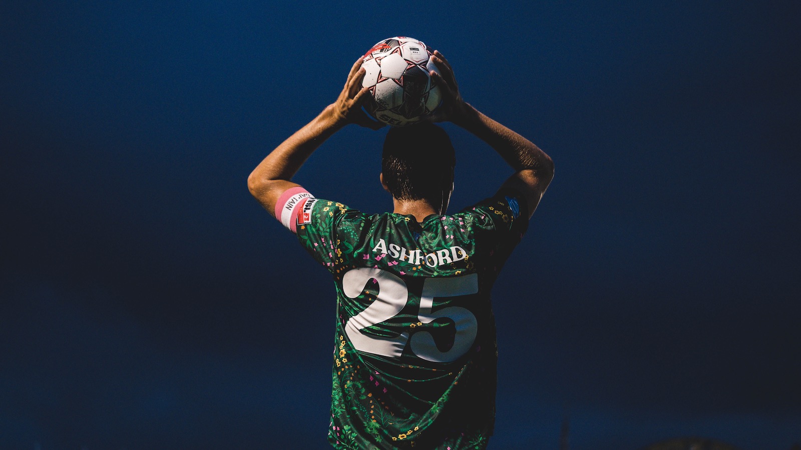 Jake Ashford, wearing a green Vermont Green FC jersey, is seen from behind against a dark sky holding the ball overhead with both hands.