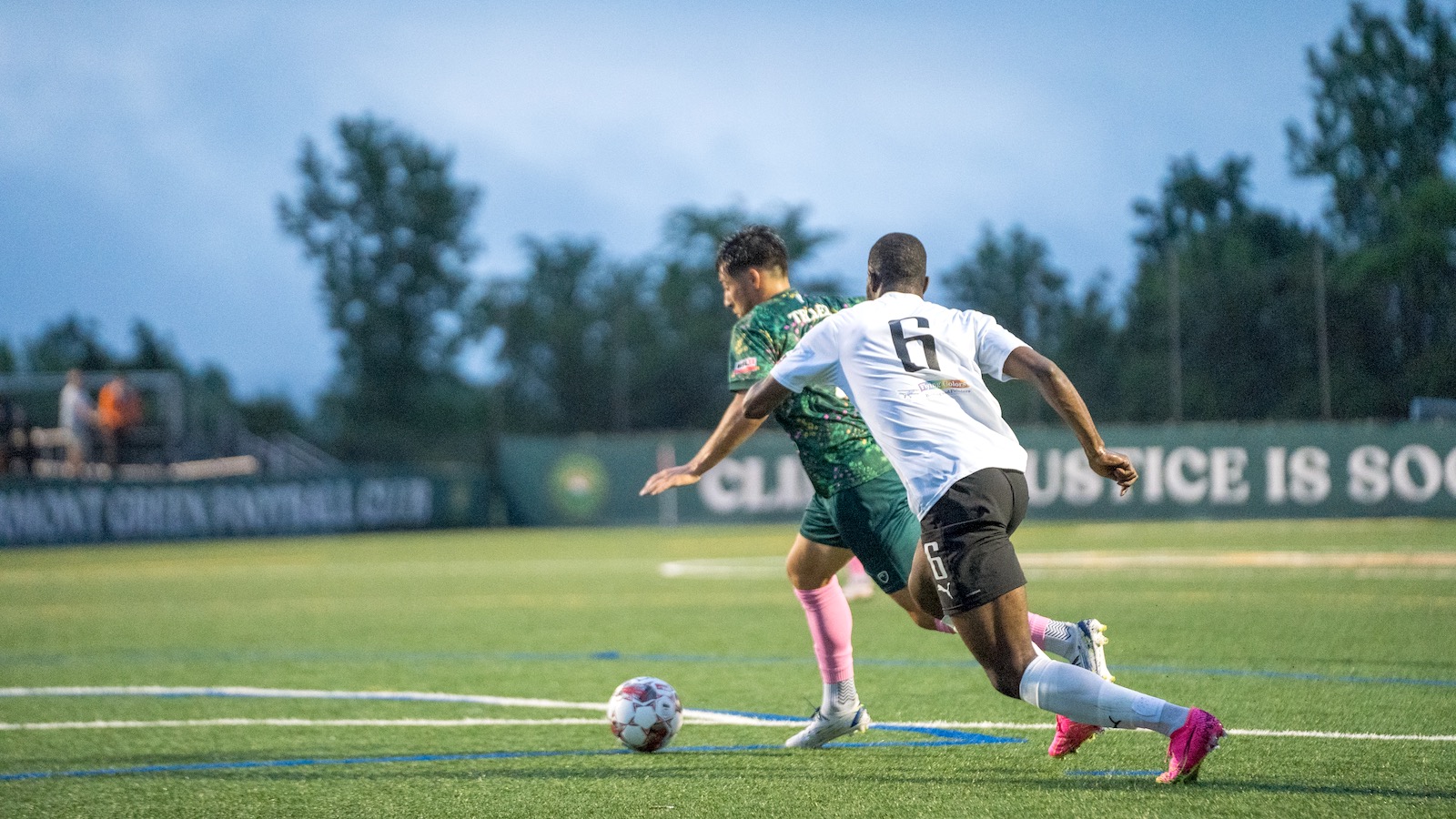Two soccer players, one in a green jersey and the other in a white jersey, chase the ball over the green pitch. A banner in the background reads, 