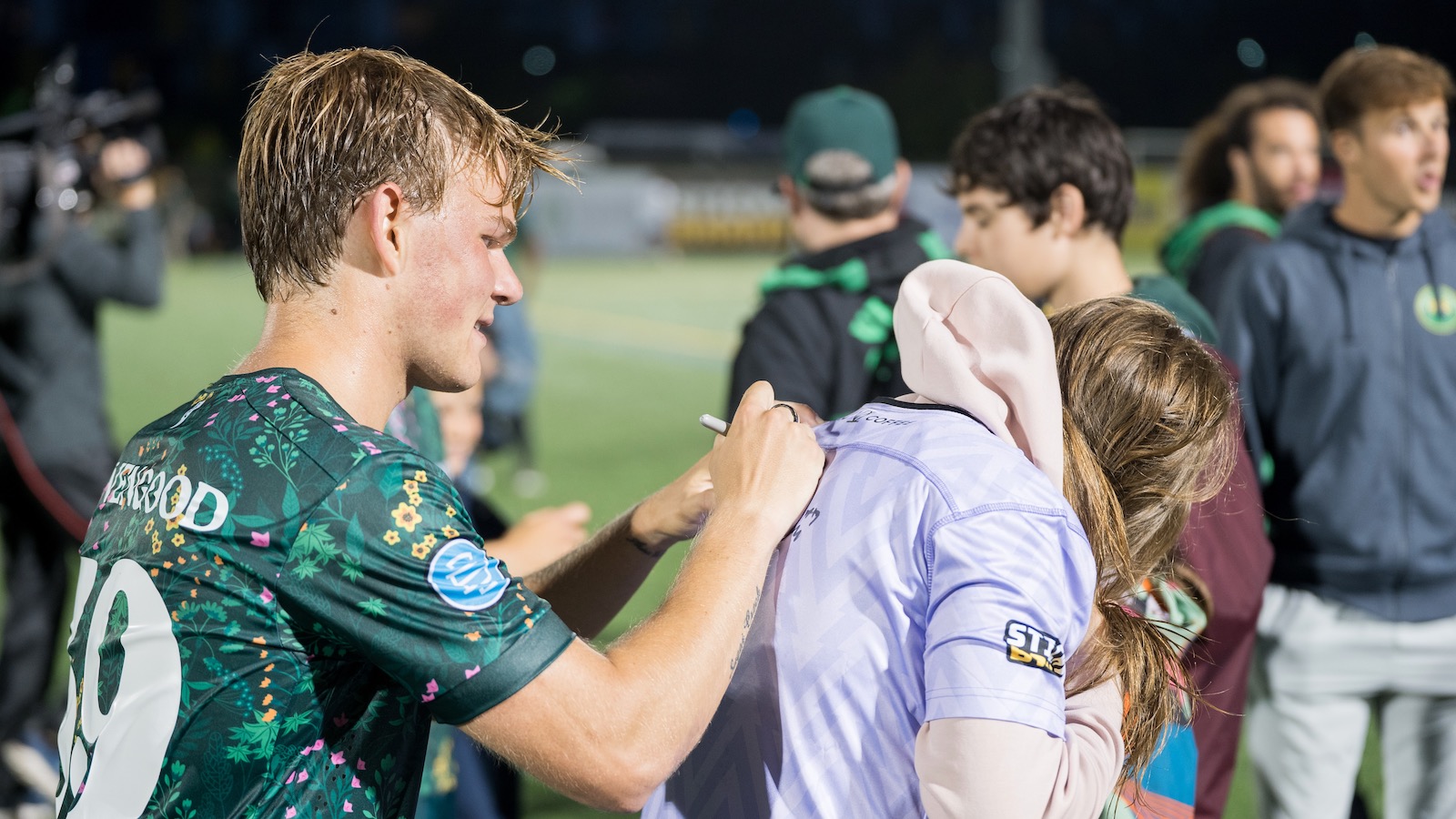 Soccer player Alexander Levengood, wearing a green Vermont Green FC jersey, signs an autograph on the back of a female fan wearing a lavender jersey over a pink hoodie.