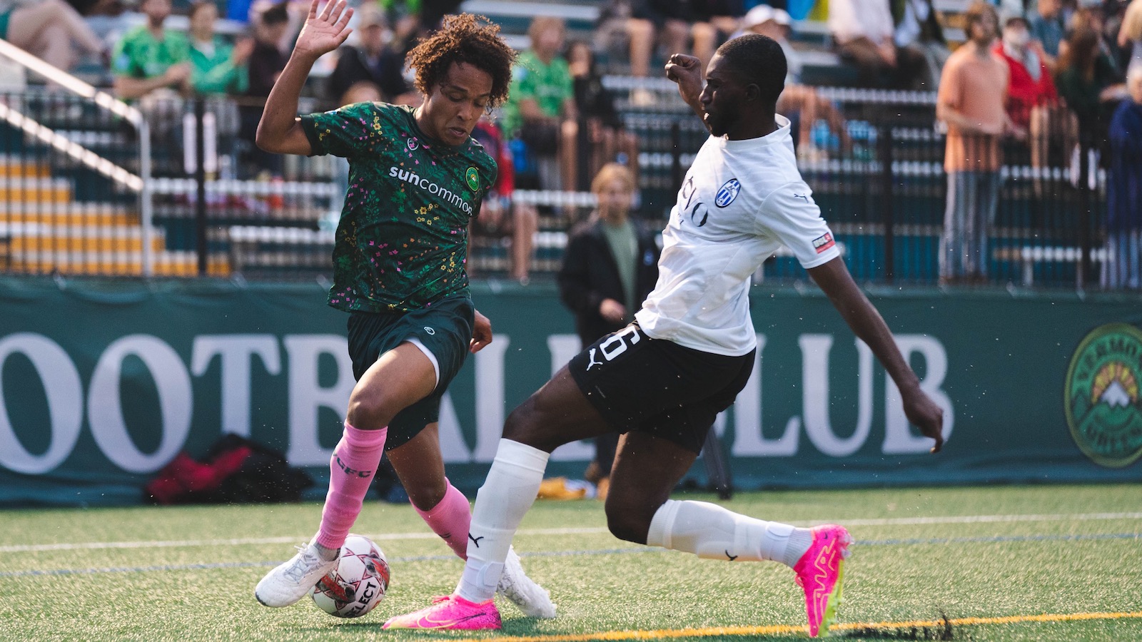 A player in a green Vermont Green FC soccer jersey, kicks the ball around an opposing player in a white jersey as fans look on from the stands behind them.
