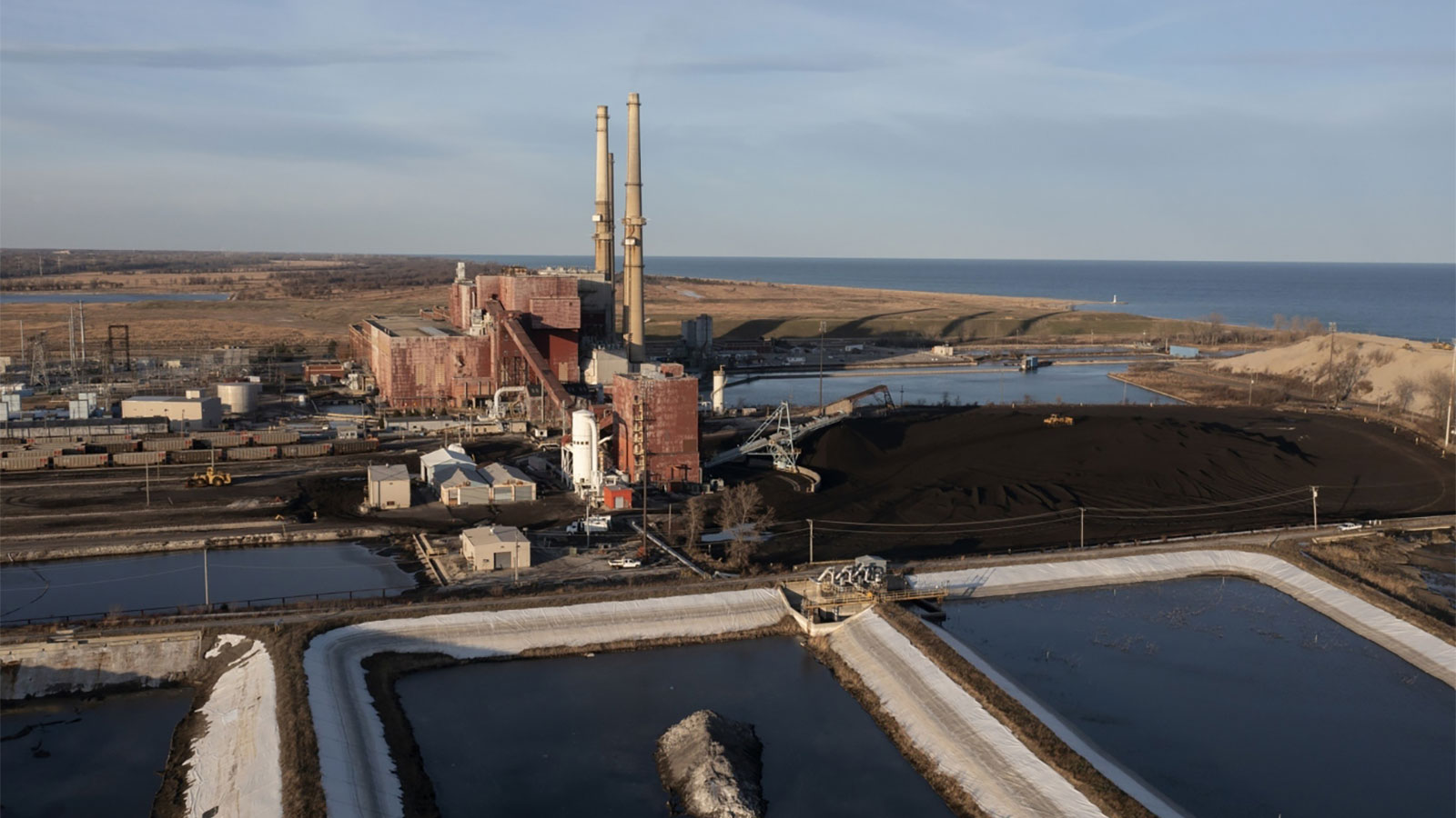ponds filled dark liquid next to smoke stacks of coal plant next to Lake Michigan