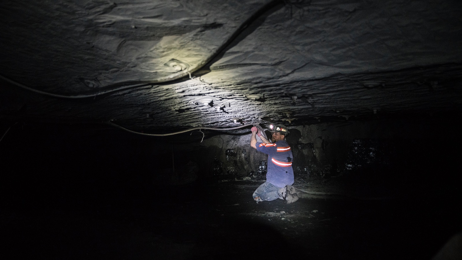 A coal miner in blue coveralls and a helmet works underground in a cramped space illuminated only by his headlamp.