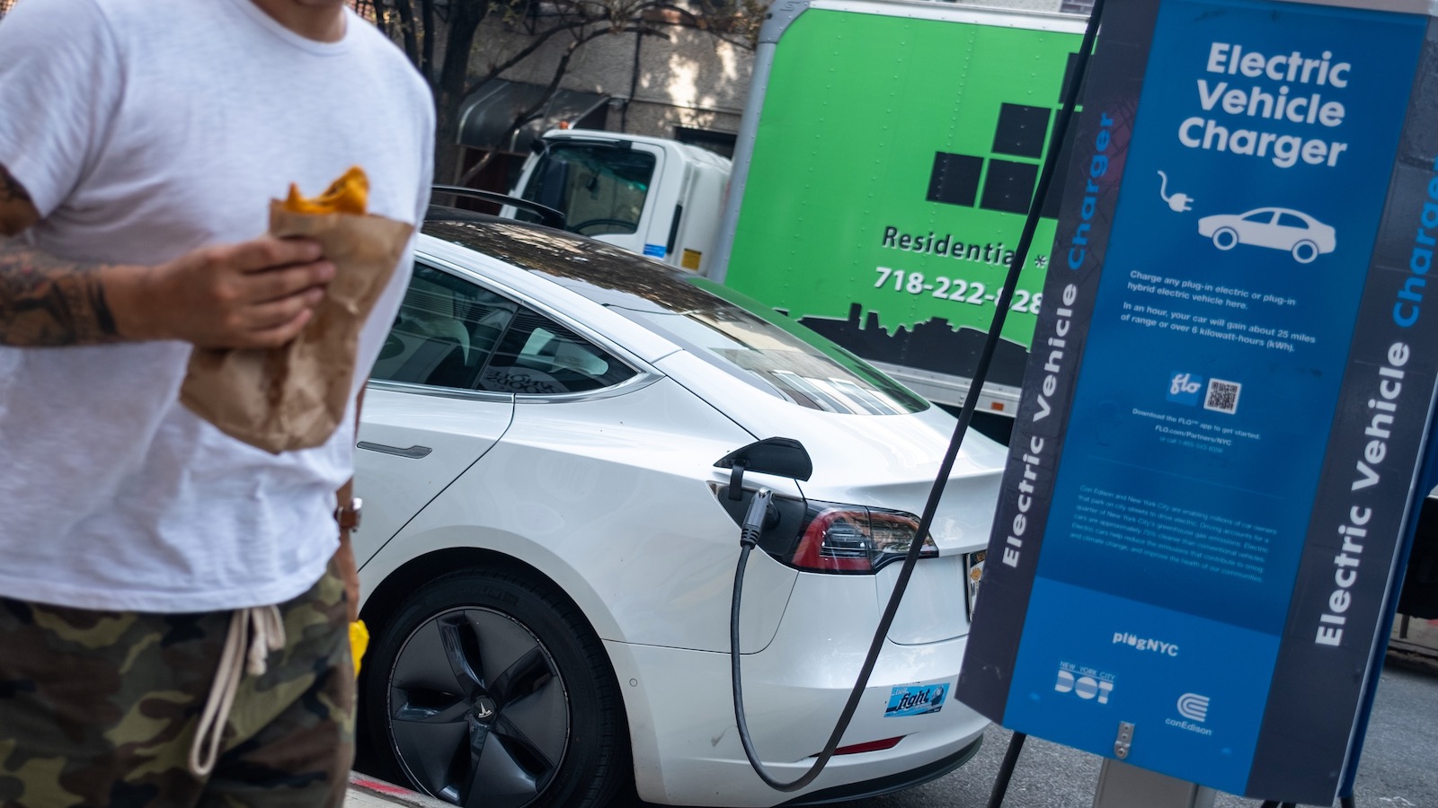 A man wearing shorts and a T-shirt and eating a burrito walks past a white Tesla Model S plugged into a public charging station in New York City.