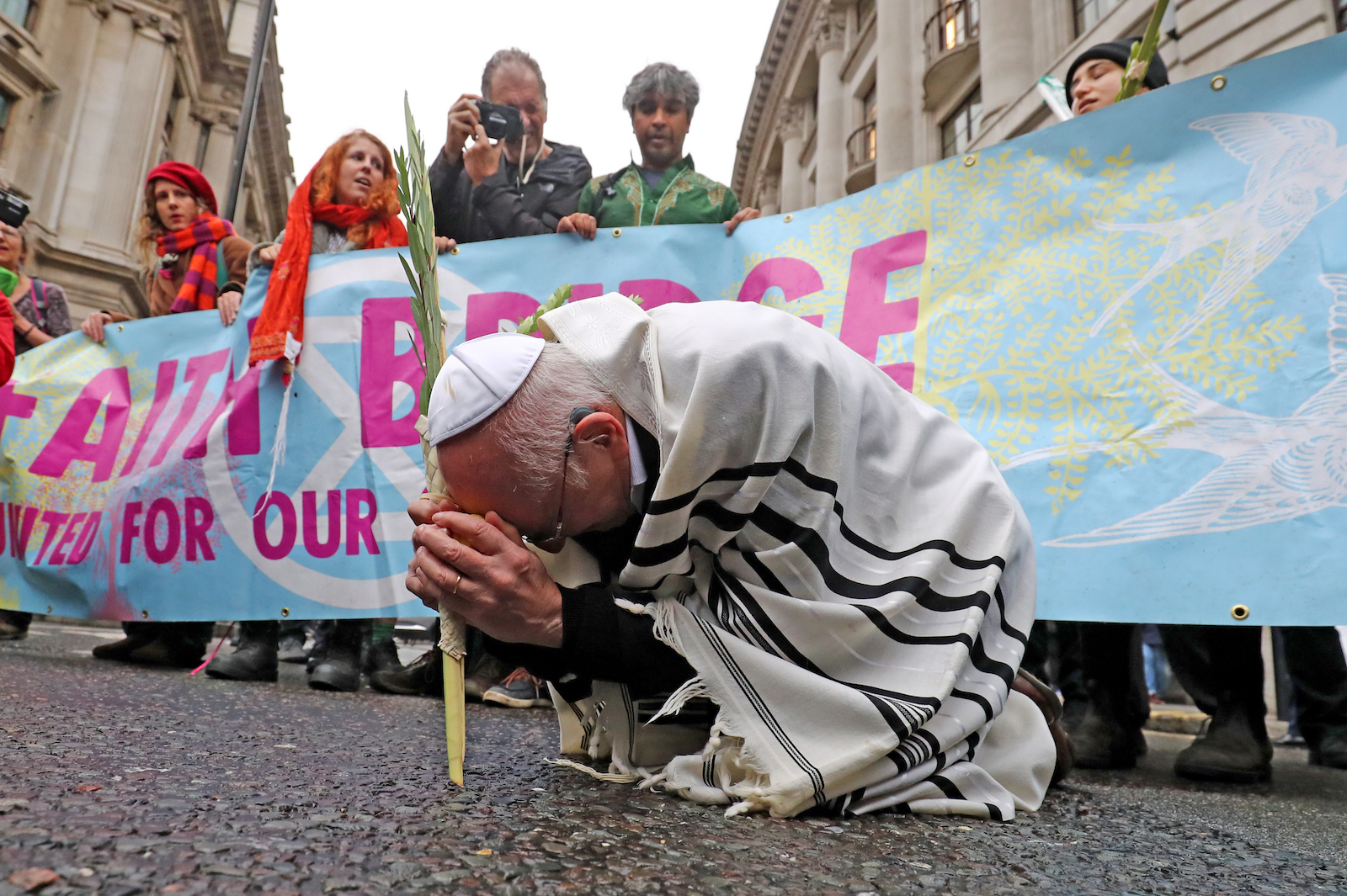 a man in traditional jewish prayer clothes kneels in front of a banner held by climate protesters