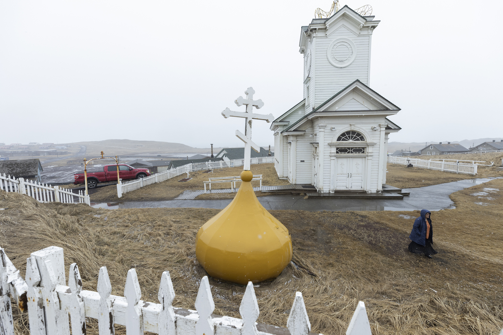A small white church surrounded by a white fence. In front is a bright yellow buoy with a cross on top