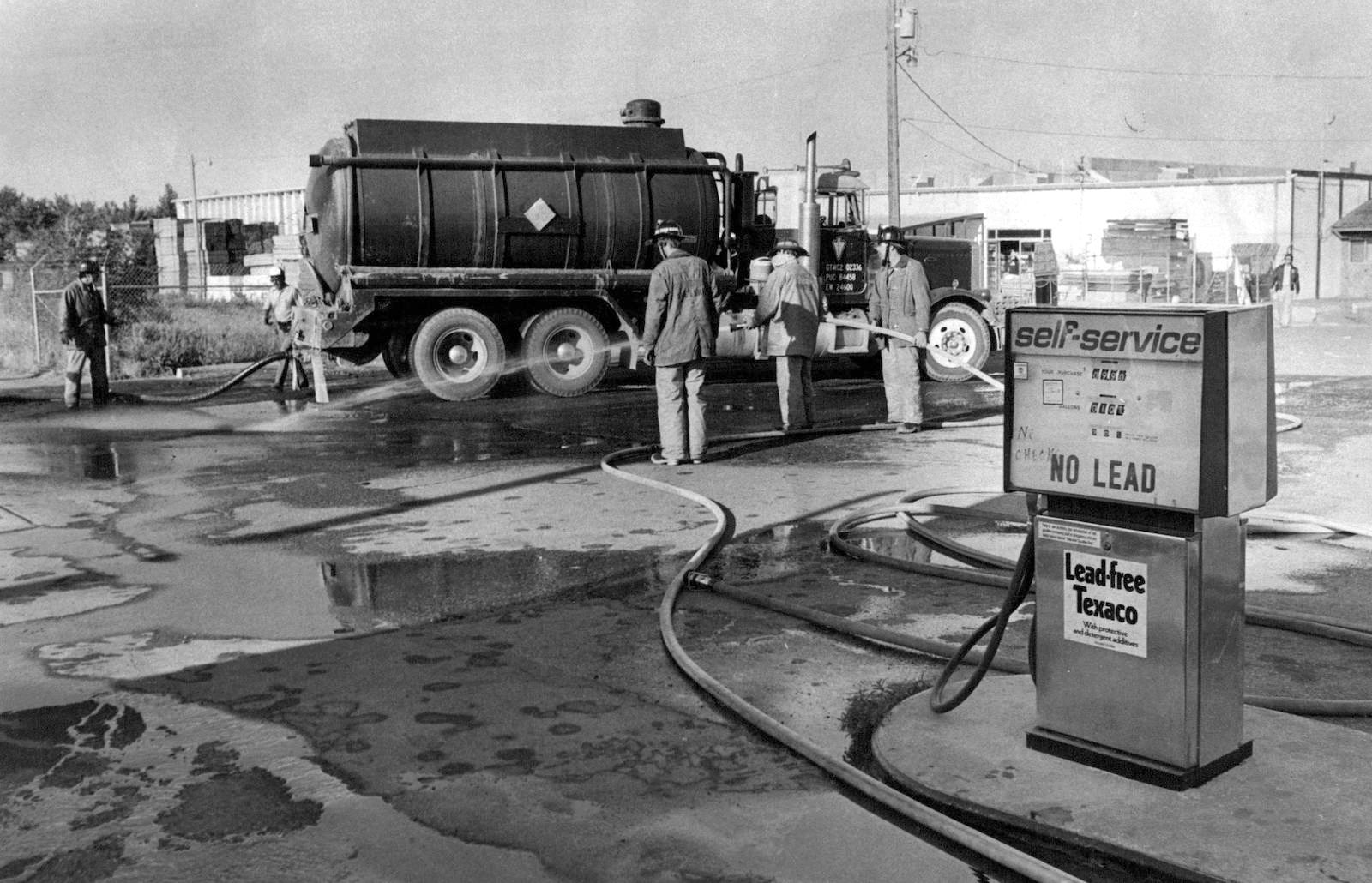 a black and white photo of firemen hosing off a gas station near a pump