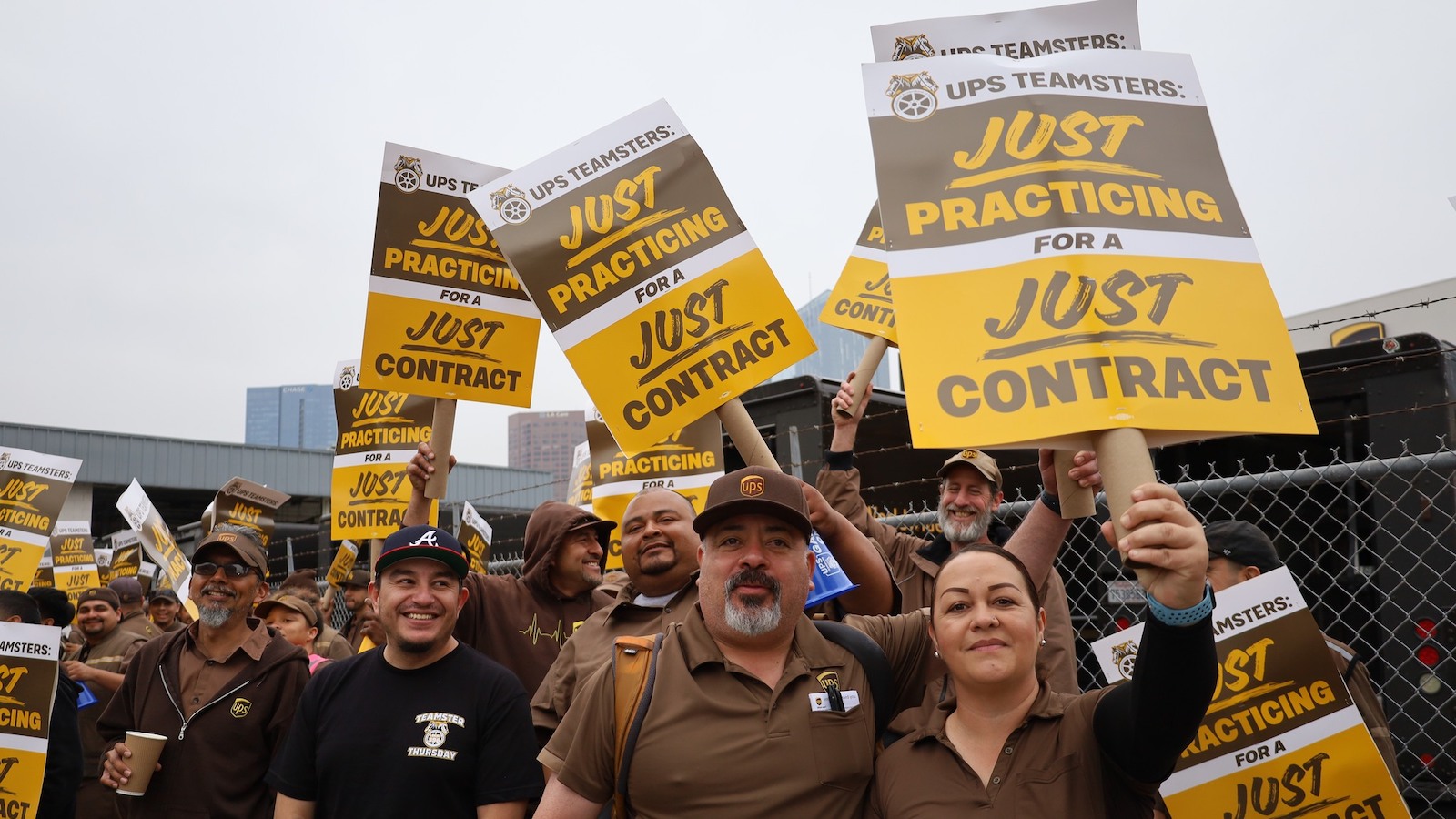 UPS workers in brown uniforms standing outside a warehouse, holding signs that say "Just Practicing for a Just Contract"