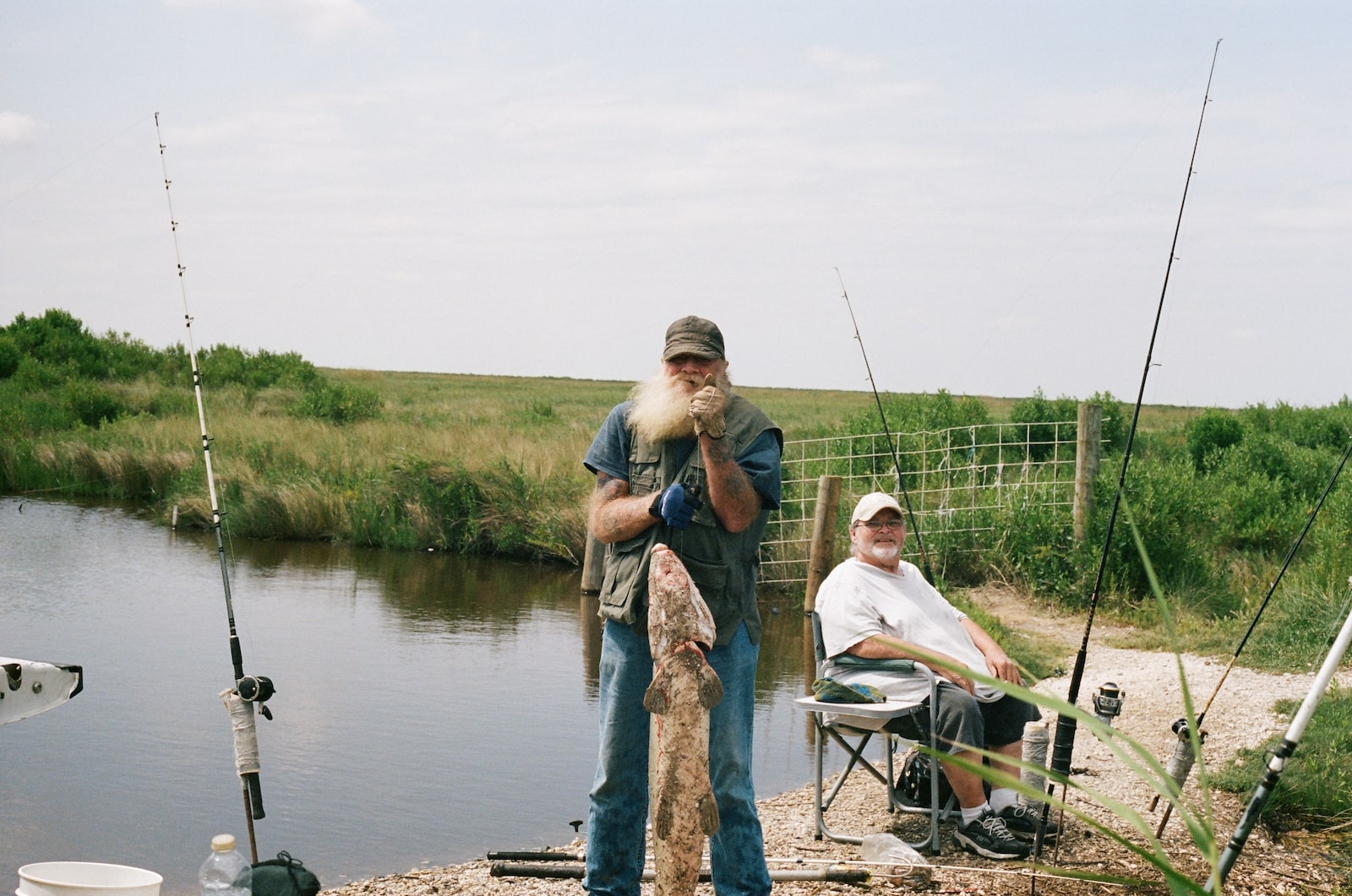 a man with a beard holds a large fish while another man sits in a chair behind him near a body of marshy water