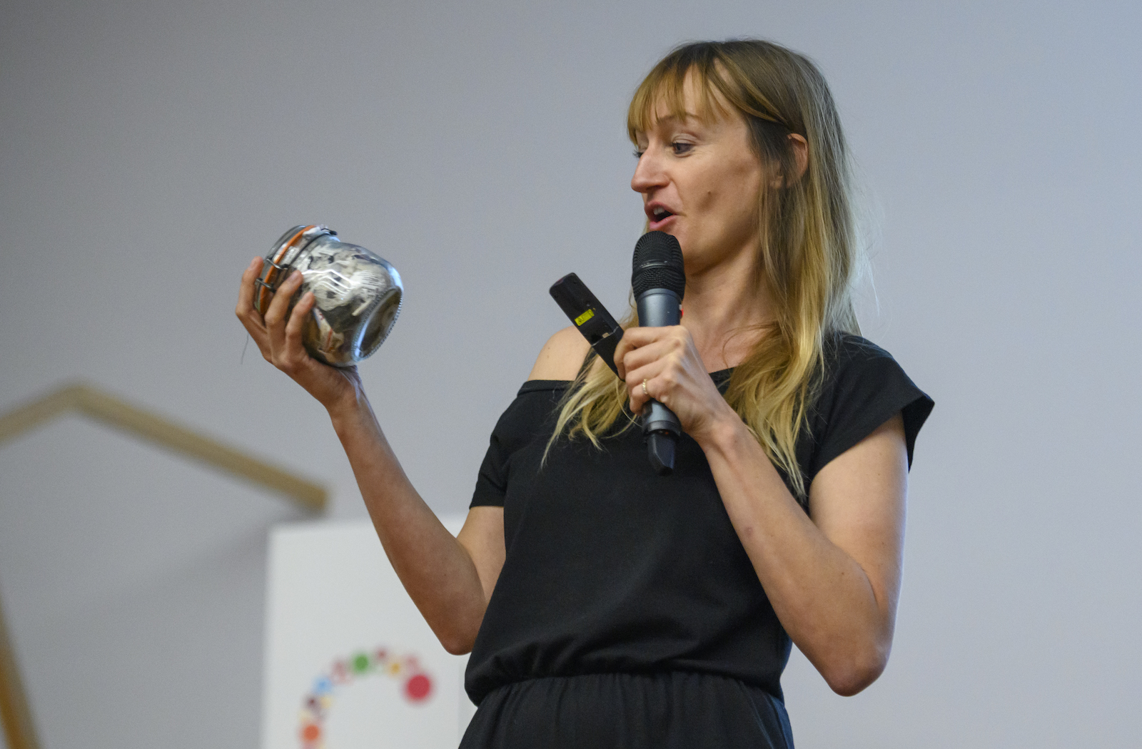 a woman in a black dress holds a microphone in one hand and a ball jar full of trash in the other