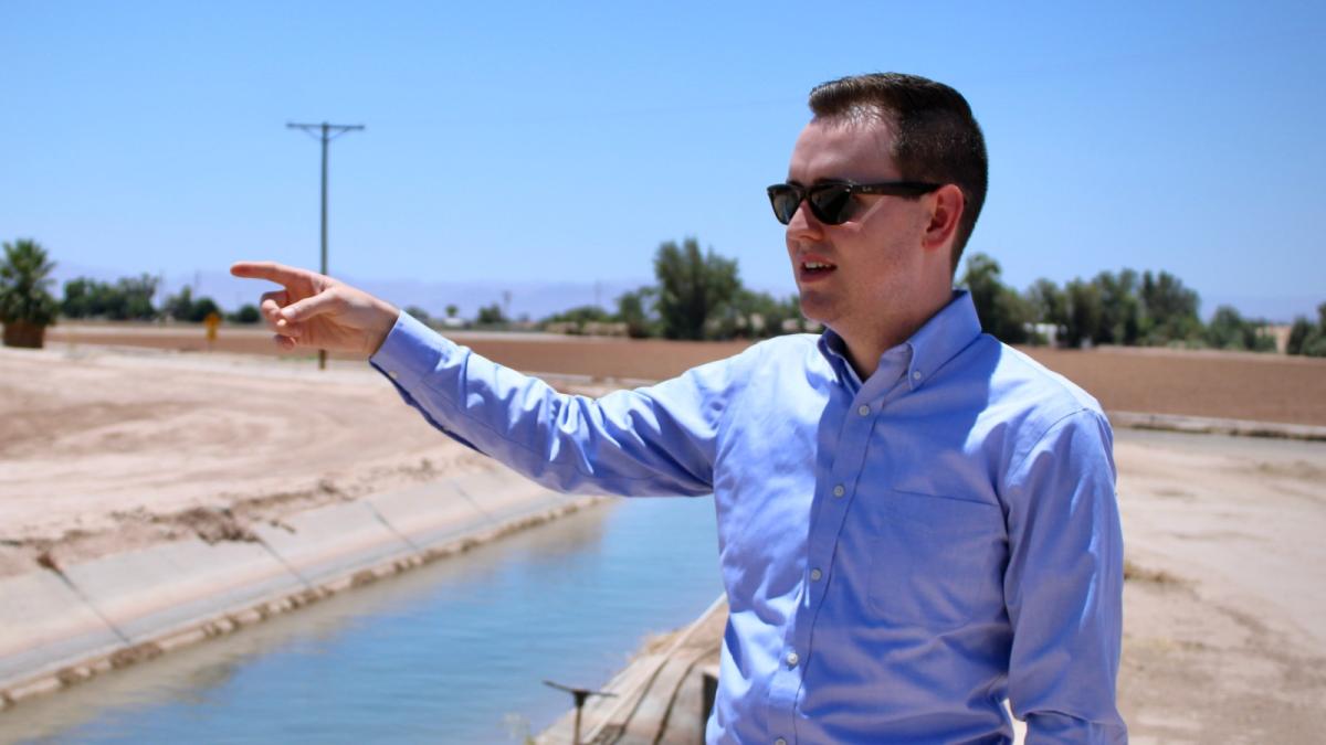 A man in black sunglasses and a blue button-down shirt points while standing next ot a canal.
