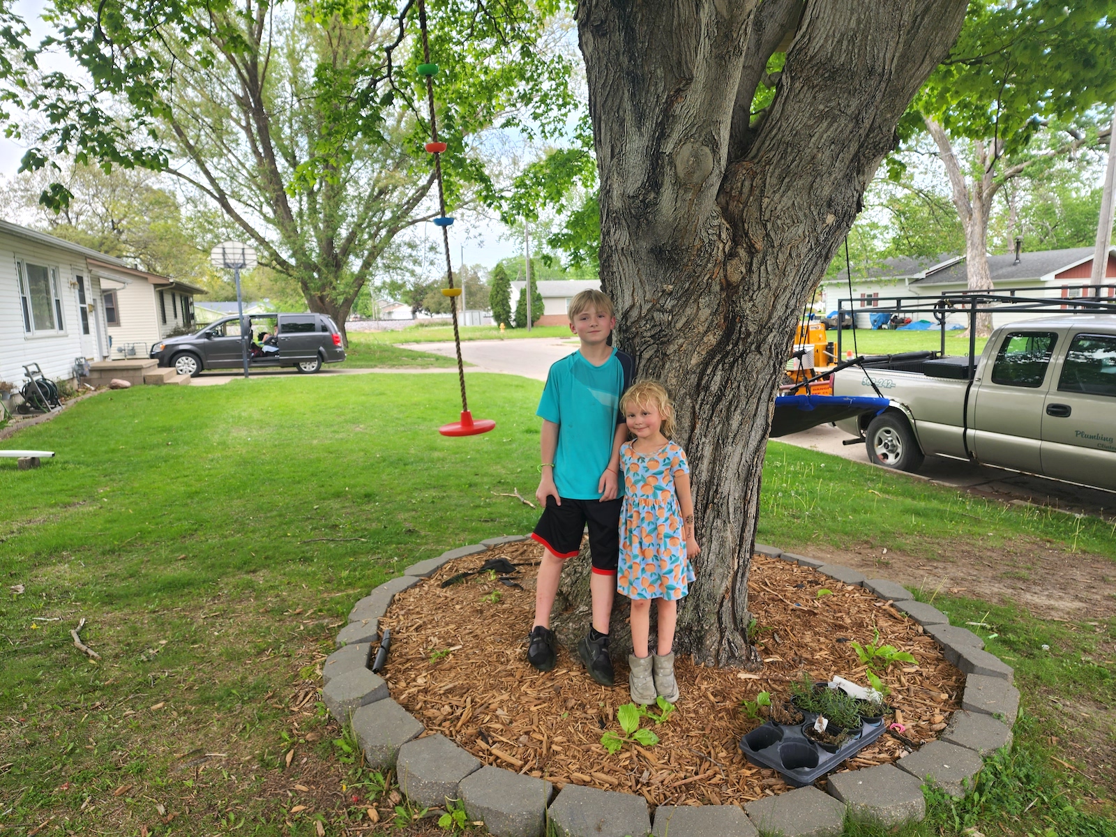two kids stand near a tree in a green yard