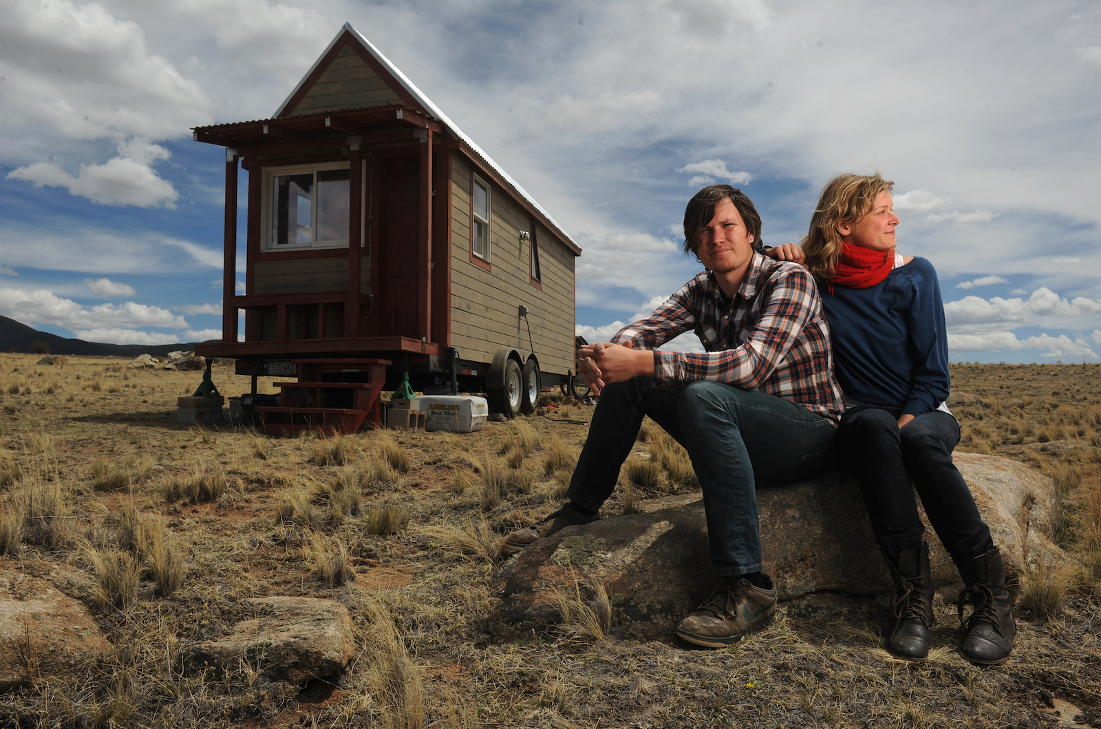 a man and a woman sit in a field in front of a tiny home