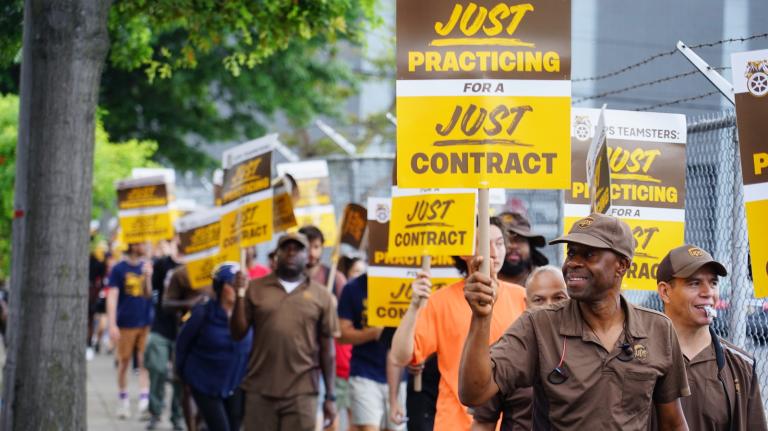 A line of UPS workers in brown uniforms walking along a sidewalk, holding signs that say 