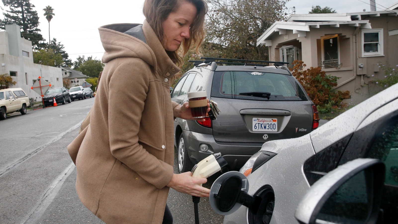 A woman in a tan coat plugs a charger into her electric vehicle, which is parked on the street outside her home in Berkeley, California
