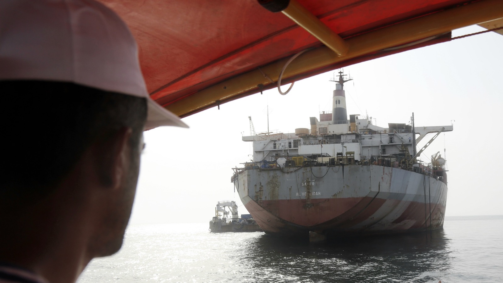 A man in a ballcap stands beneath the awning on the deck of a small ship approaching the FSO Safer, a decaying oil tanker on the Red Sea.