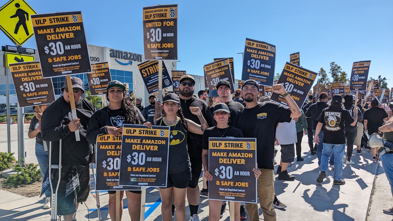 A crowd of amazon delivery drivers standing in front of a warehouse holding signs that say 