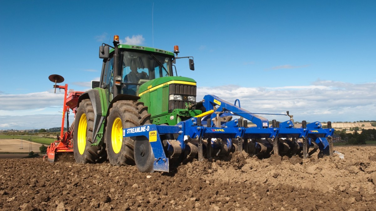 A green tractor with a blue grill runs through a field of soil under a blue sky.
