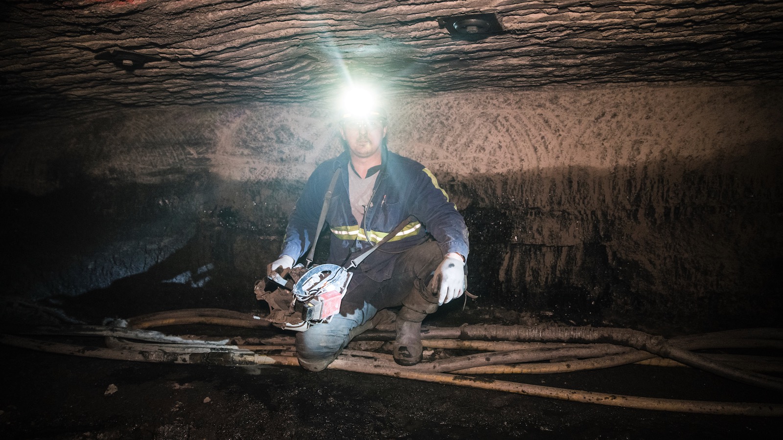 A coal miner in blue coveralls and a headlamp looks directly at the camera while crouching in the cramped confines of a mine shaft.