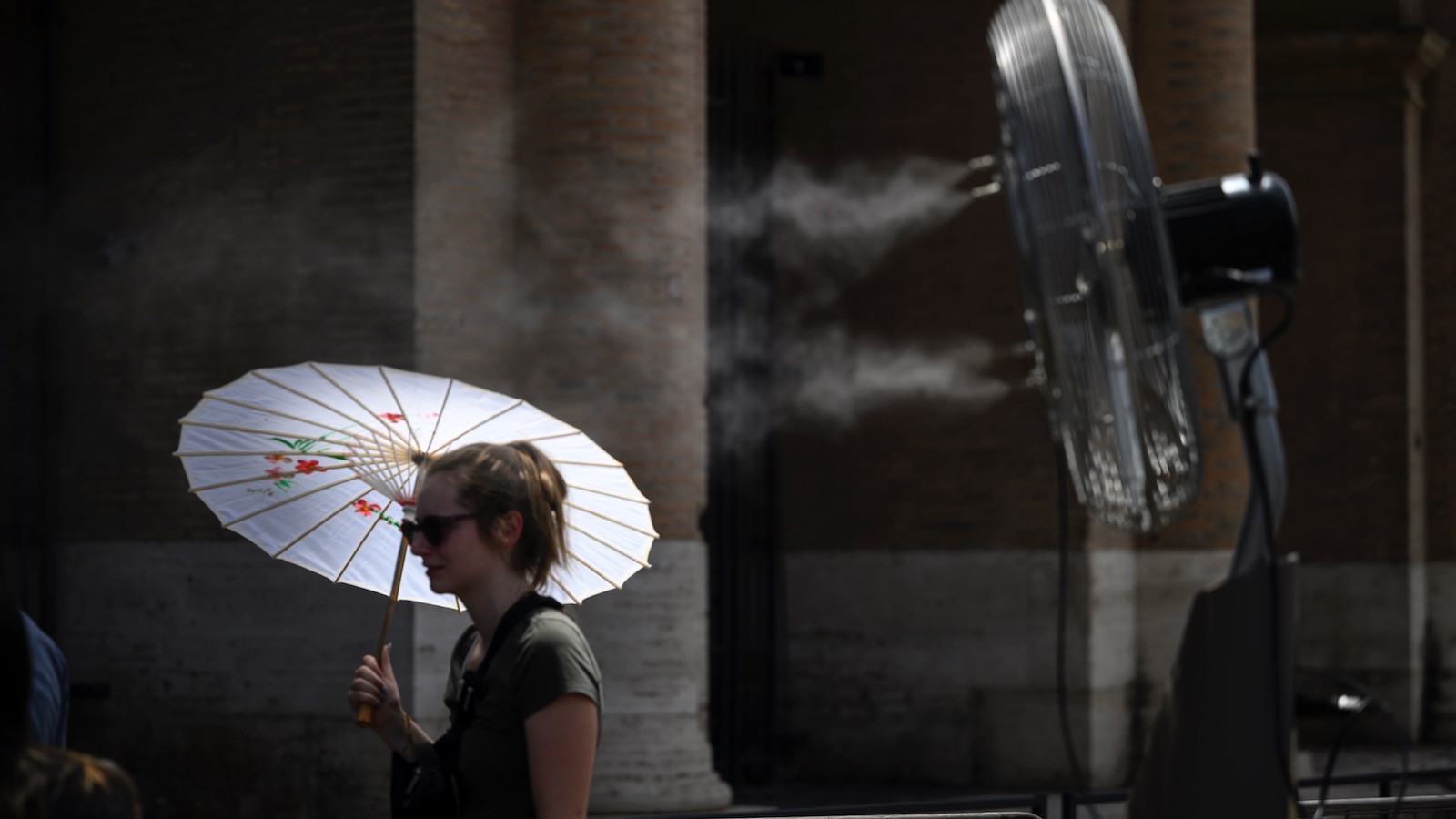 A young woman carrying a parasol walks past an electric fan blowing cool mist over a sidewalk in Rome as temperatures reach 44 degrees Celsius (111 degrees Fahrenheit).