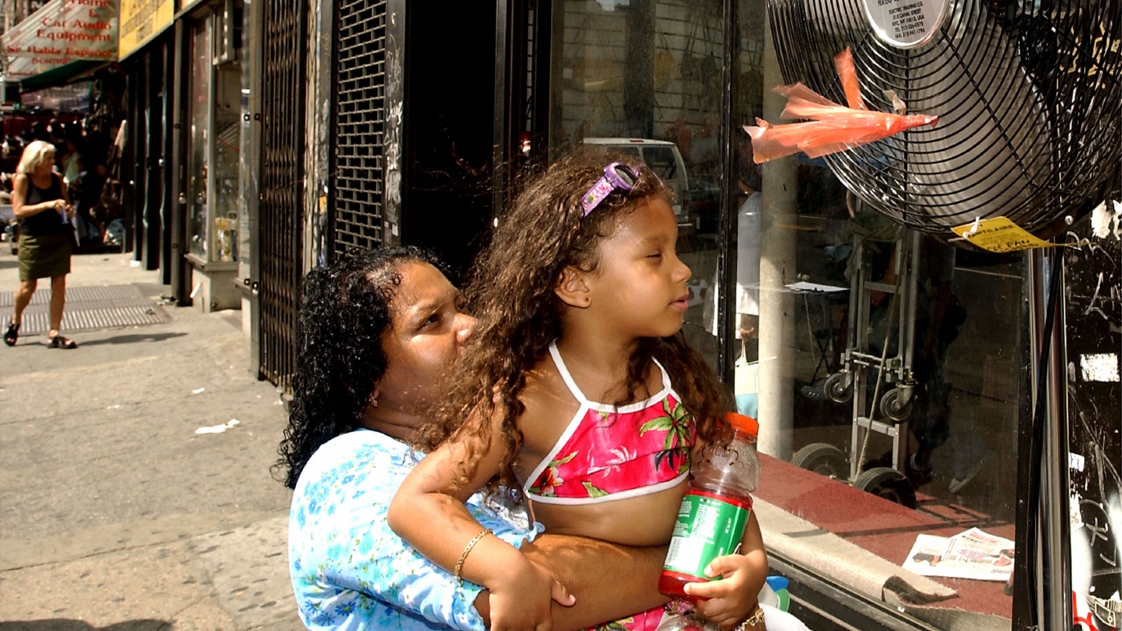 A mother holds a daughter in front of a fan on a street in New York City during a heat wave.