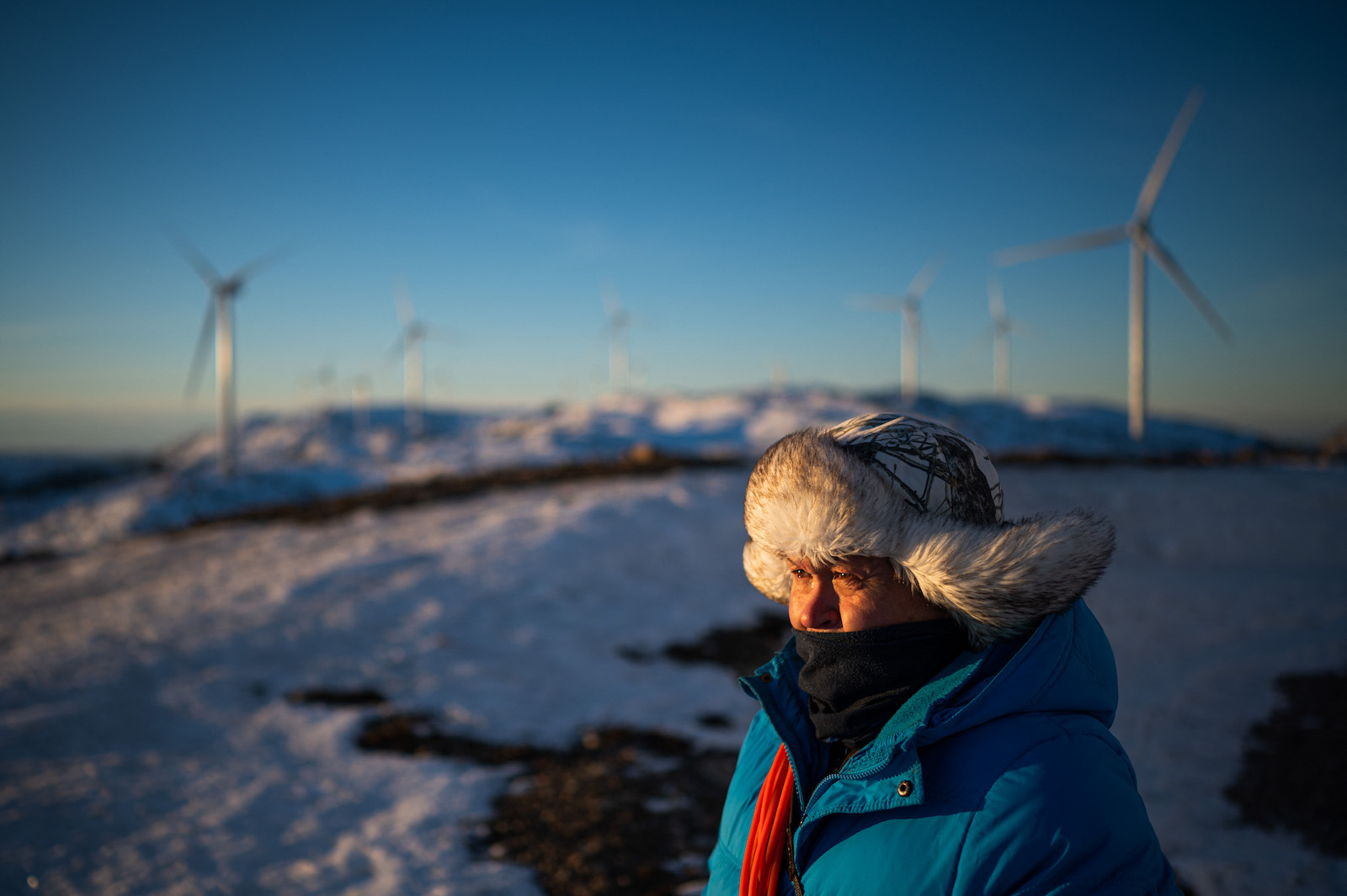 a man in a furry hat looks out with wind turbines in the distance