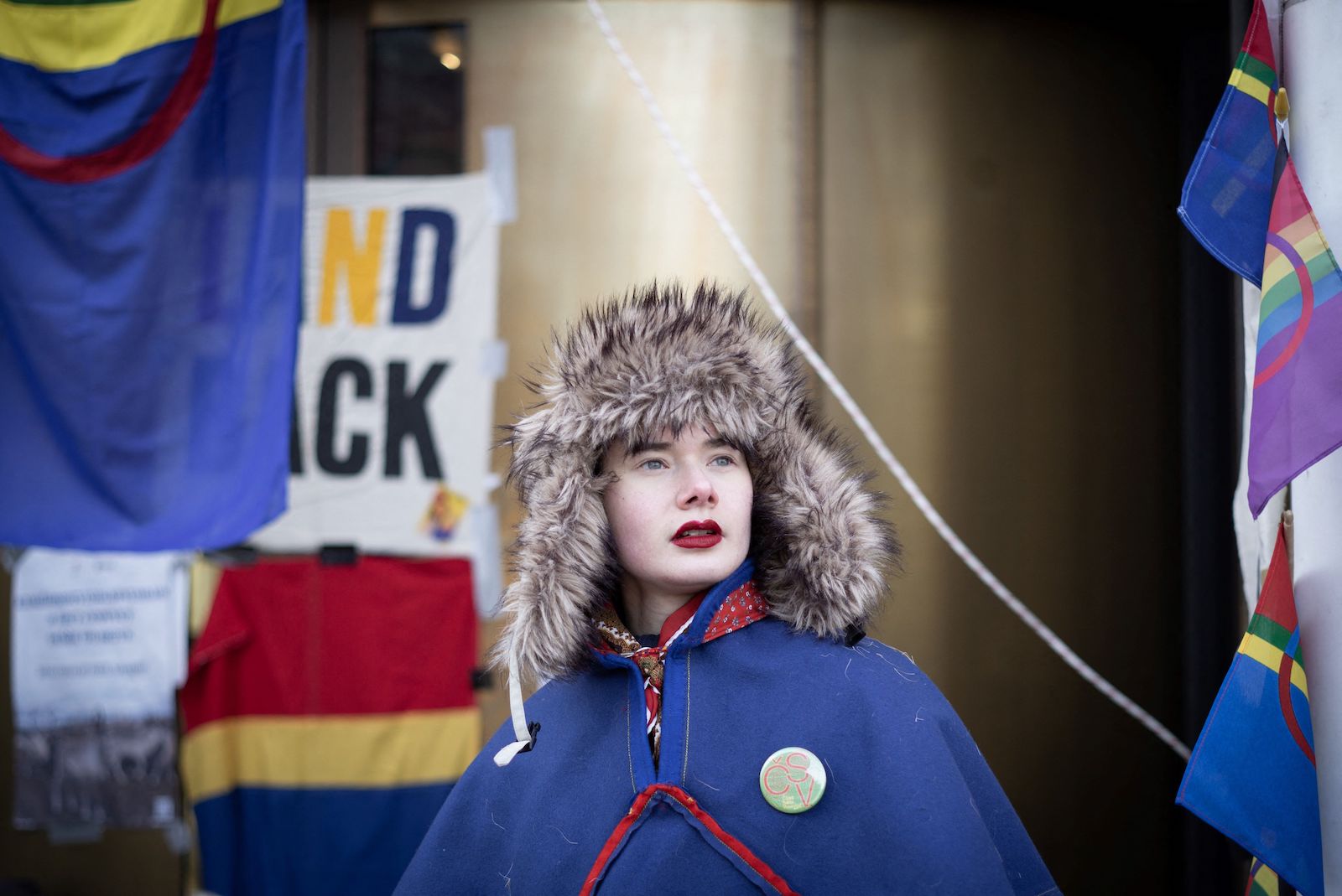 A young woman in a fur hat and dark blue cloak stands in front of a sign on the back of the earth