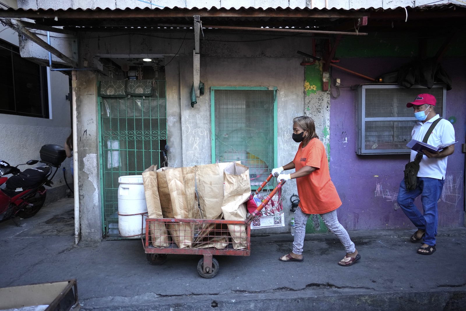 a woman in an orange shirt pushes a cart full of paper bags of trash through a town
