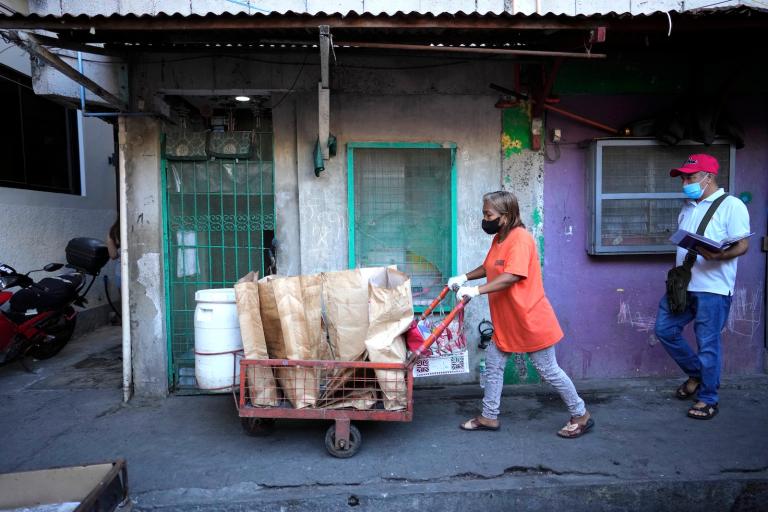 a woman in an orange shirt pushes a cart full of paper bags of trash through a town