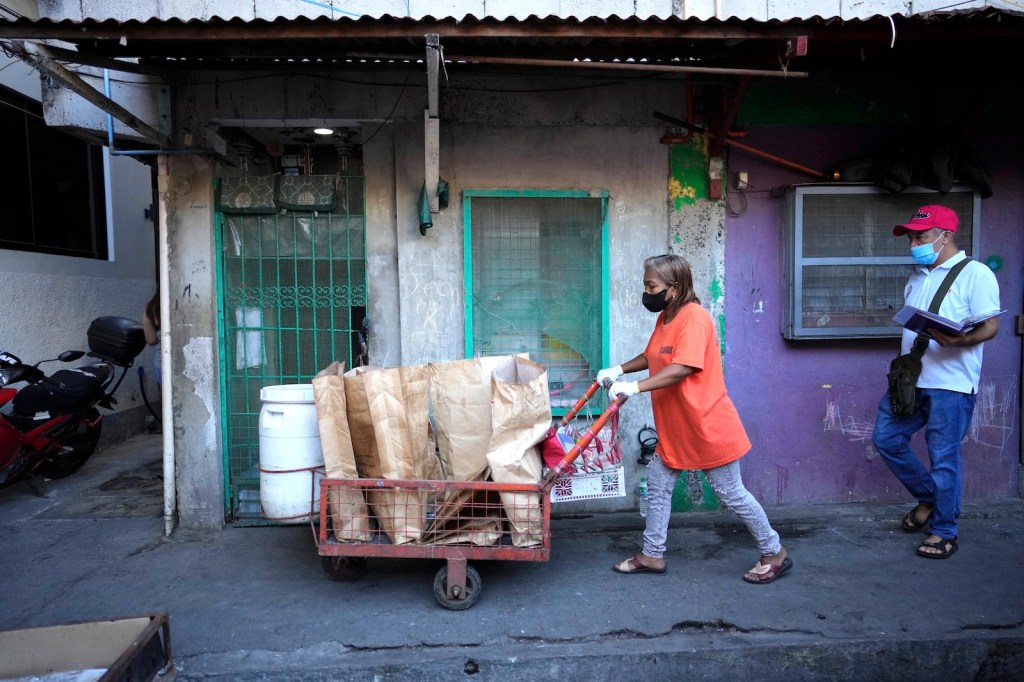 a woman in an orange shirt pushes a cart full of paper bags of trash through a town