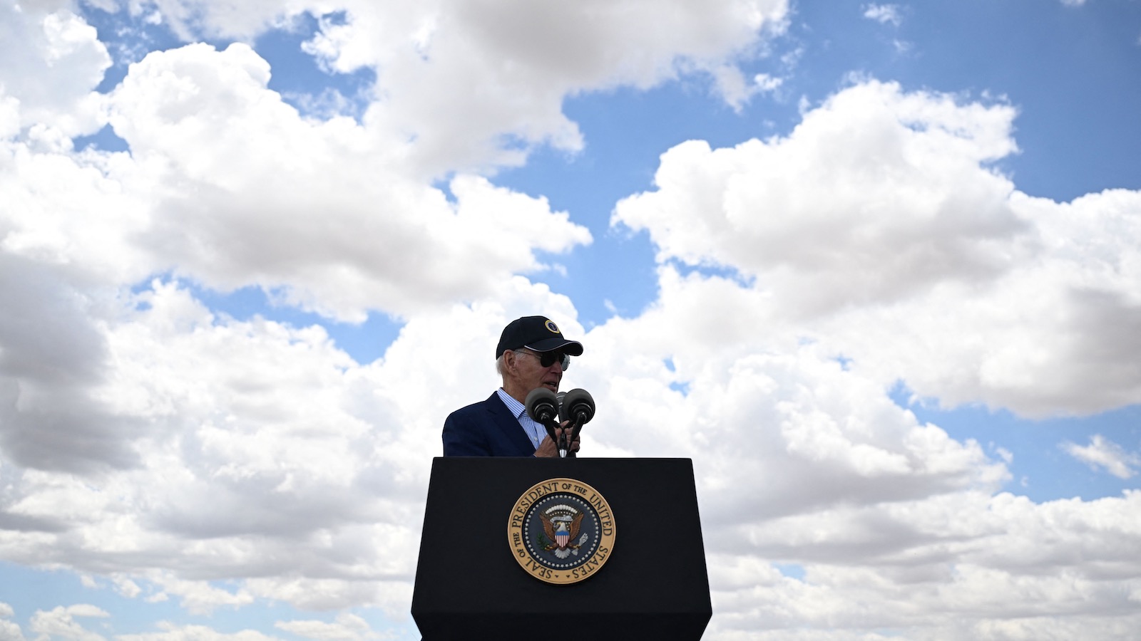 President Joe Biden, wearing a ball cap and sunglasses, speaks at a lectern outdoors.