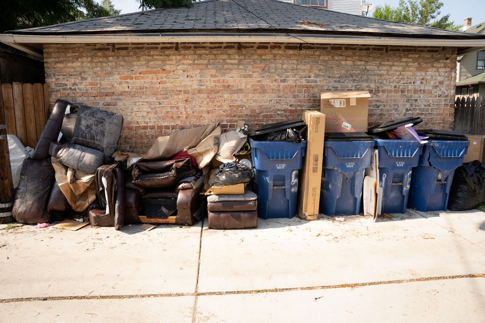 Trash bins and damaged furniture sit outside of a house.
