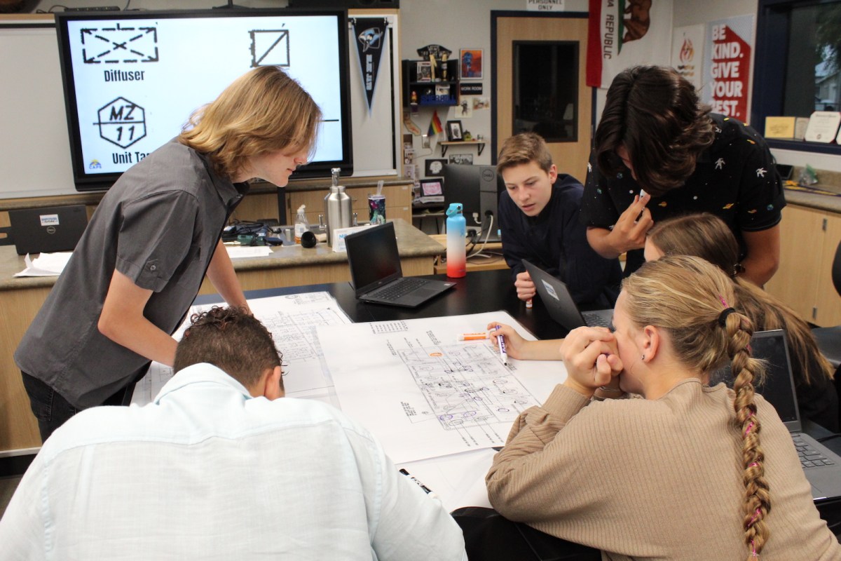 a group of teens gather around a table in a classroom