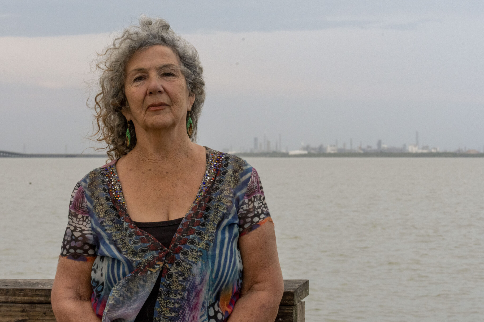 A woman with curly gray hair and a blue blouse stands in front of a bay.