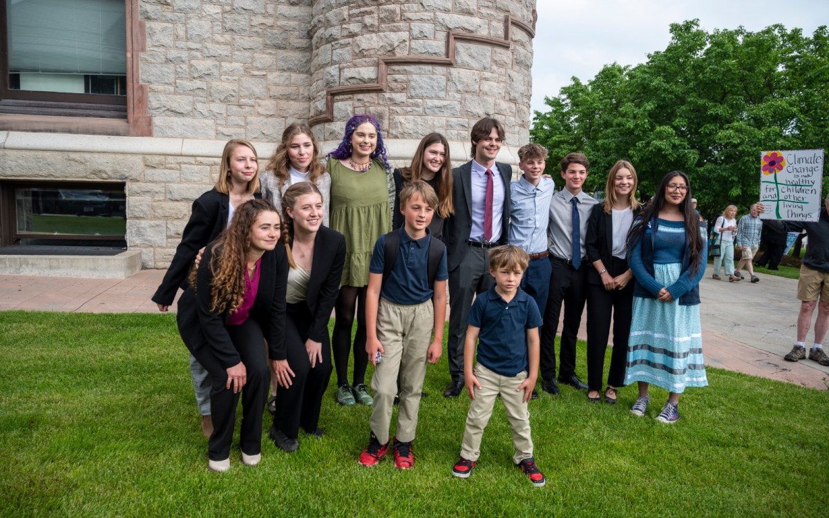 Thirteen young people cluster together on a green lawn in front of a stone building, smiling for the camera.