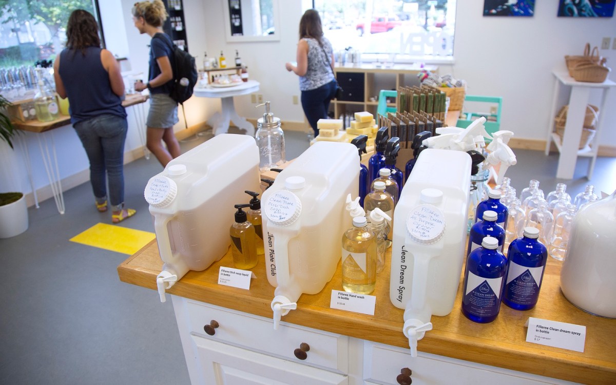 An assortment of small, fillable bottles and large plastic dispensers filled with liquid soaps sit on top of a wooden counter. In the background, three women peruse products.