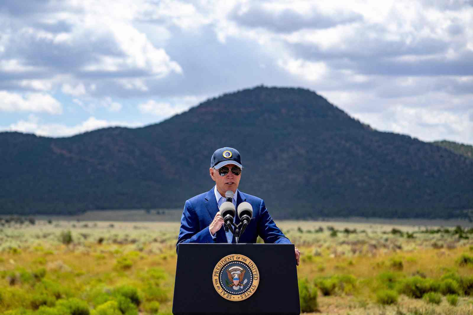 a man in a hat and suit stands in front of a podium with the presidential seal. Behind him, a beautiful field and mountain