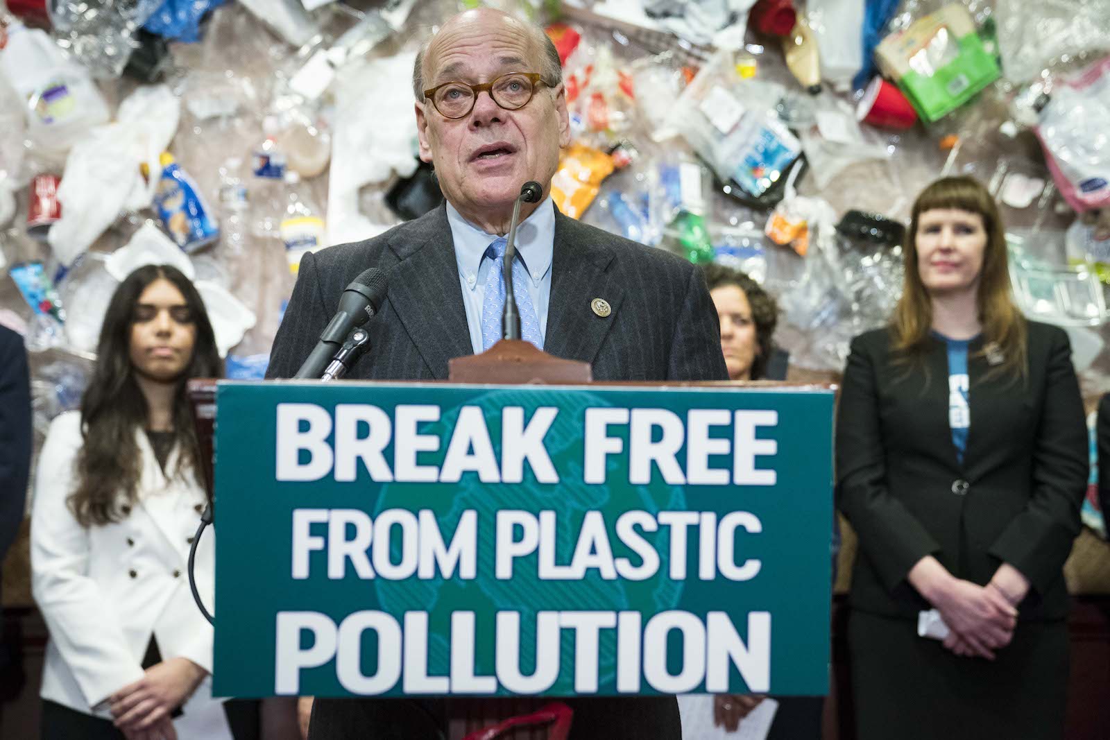 a man in a suit stands in front of a pile of plastic garbage at a podium that says break free from plastic