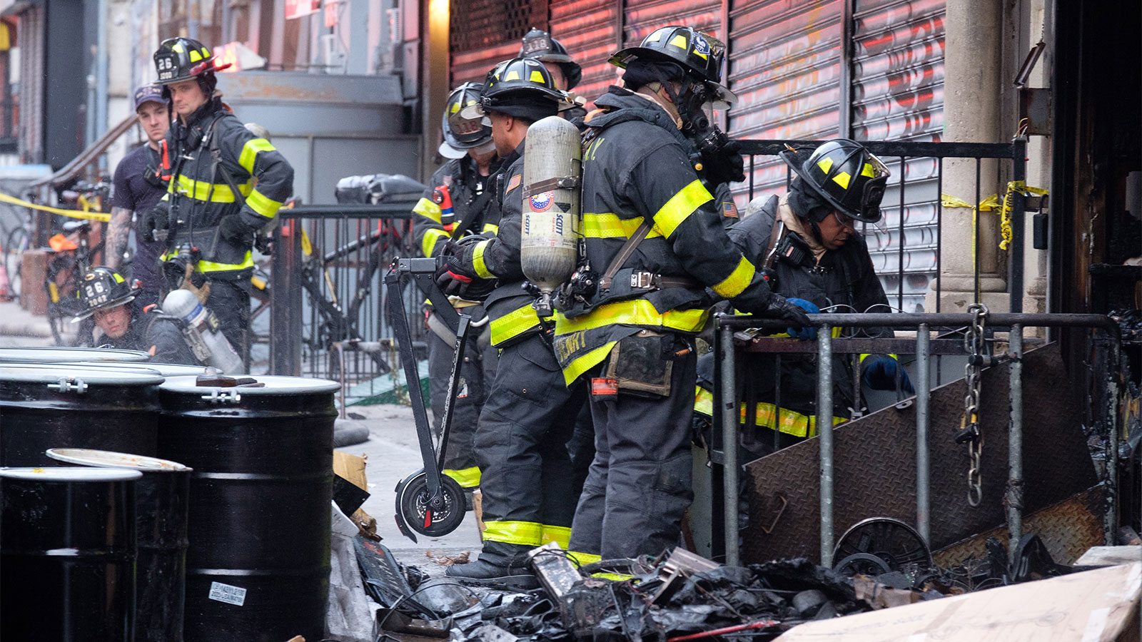 Several firefighters wearing helmets and black fireproof gear stand on a sidewalk in between a shuttered storefront, some junk, and some cylindrical bins