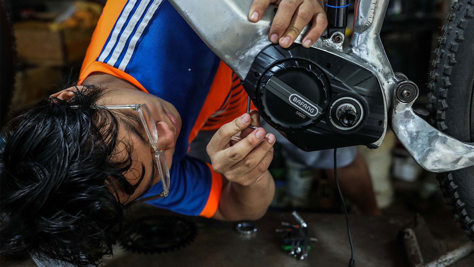 A man wearing glasses leans over to work on the frame of an e-bike with a tool