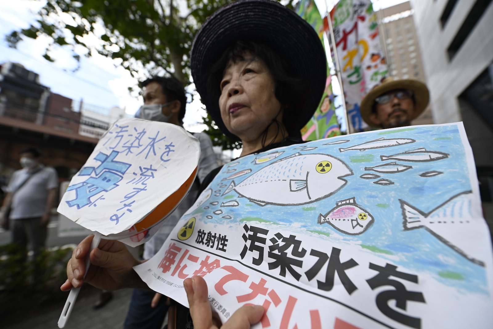 a woman stands amid a group of protesters. She is holding a sign with a drawing of a fish with nuclear symbol eyes