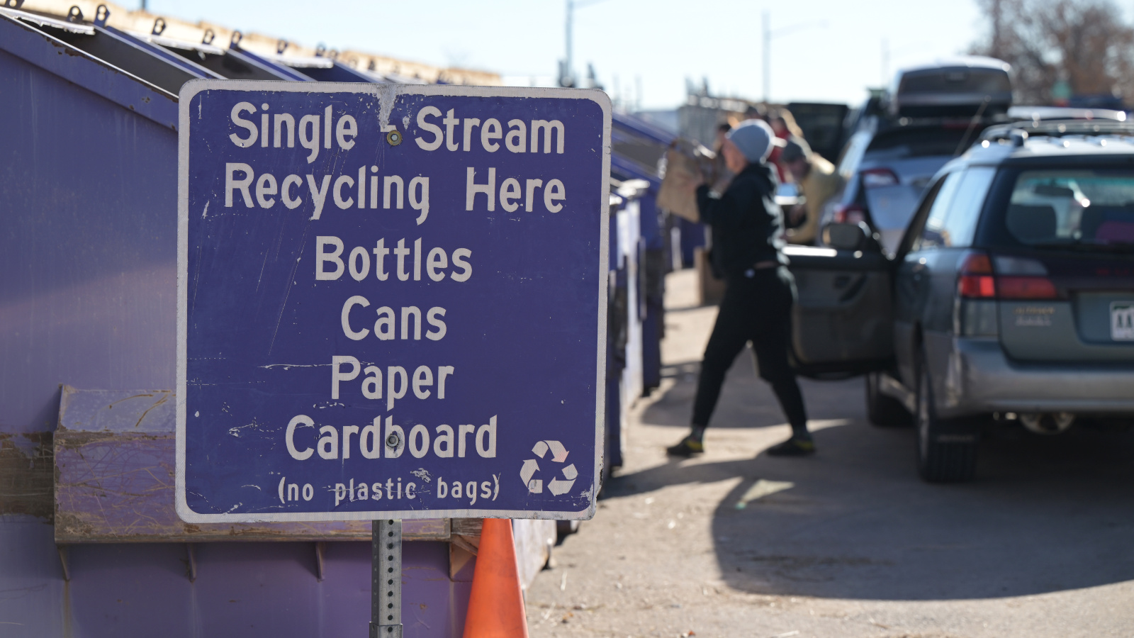 Photo of people putting recycling into dumpsters. A sign says bottles, cans, paper, and cardboard are accepted