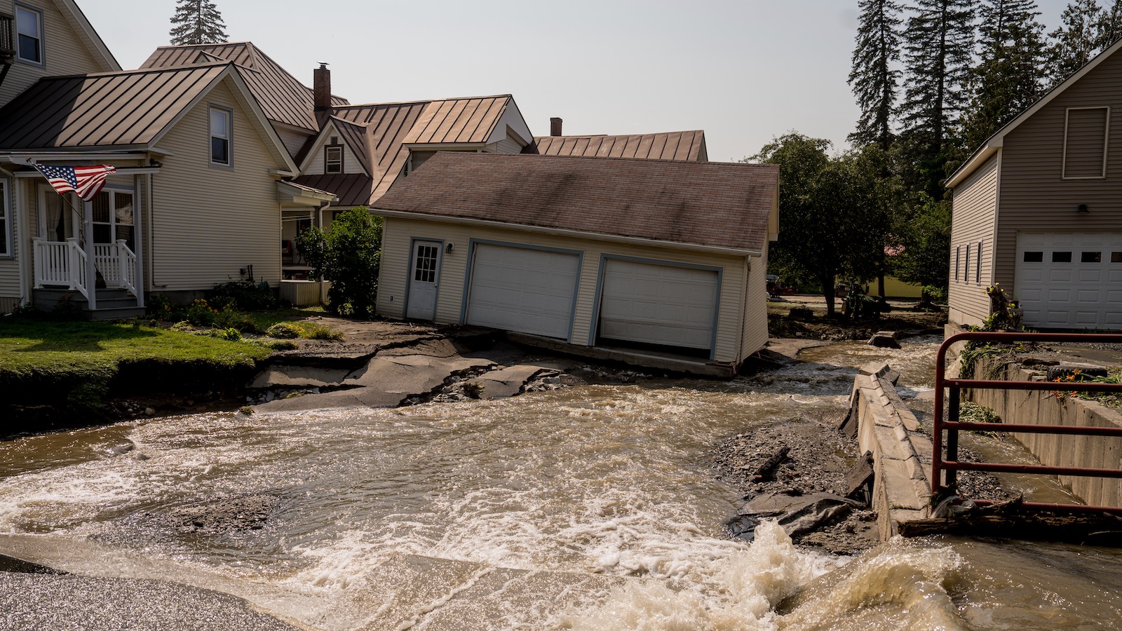 A two-car garage leans precariously on its foundation as a torrent of water flows over a crumbled and undulating driveway following a flood in Barre, Vernont.