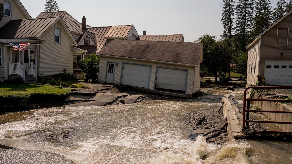 A two-car garage leans precariously on its foundation as a torrent of water flows over a crumbled and undulating driveway following a flood in Barre, Vernont.