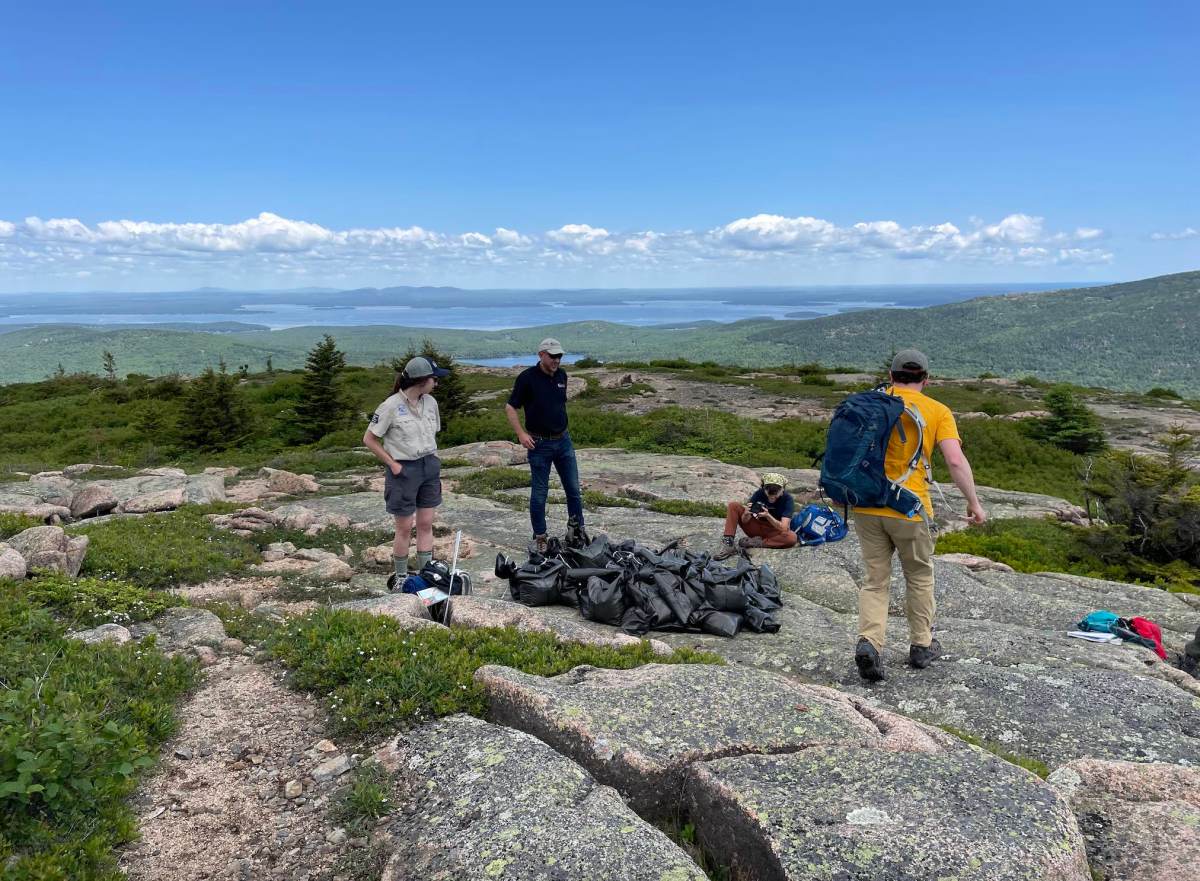 On a rocky mountain face, four people in outdoor clothing stand around a pile of black plastic bags beneath a blue sky.