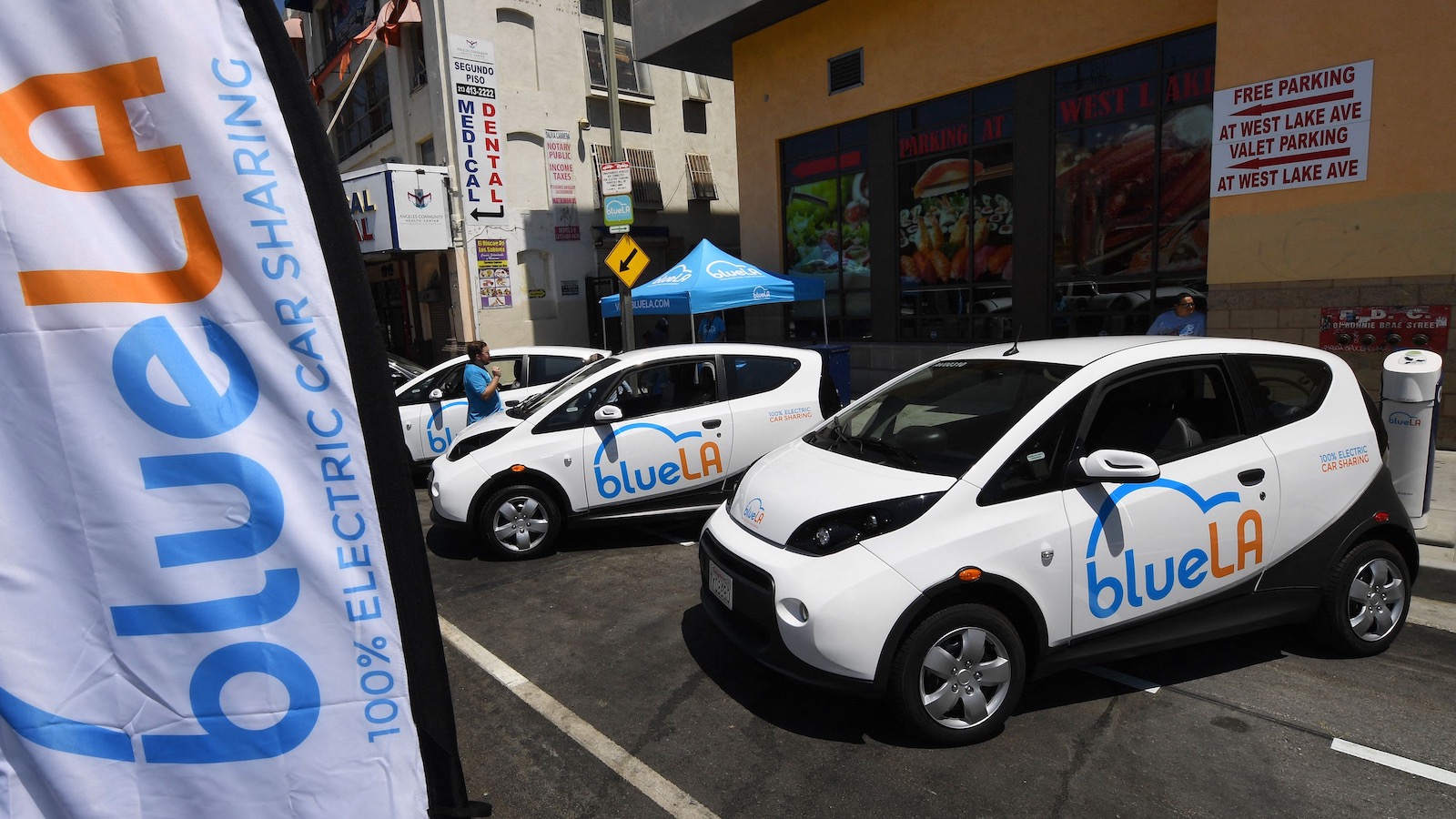 A line of three electric vehicles are parked along a street in Los Angeles with a large banner reading "Blue LA 100 percent electric car sharing" billowing in the foreground.