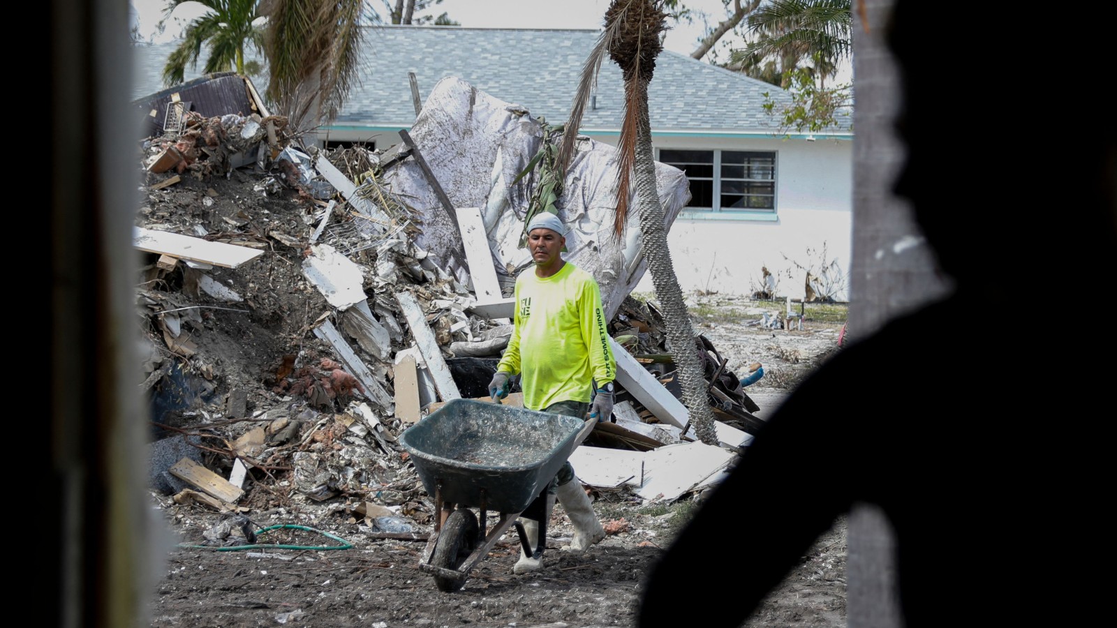 A man in a yellow shirt pushes a wheelbarrow amid mounds of debris as another man watches on.