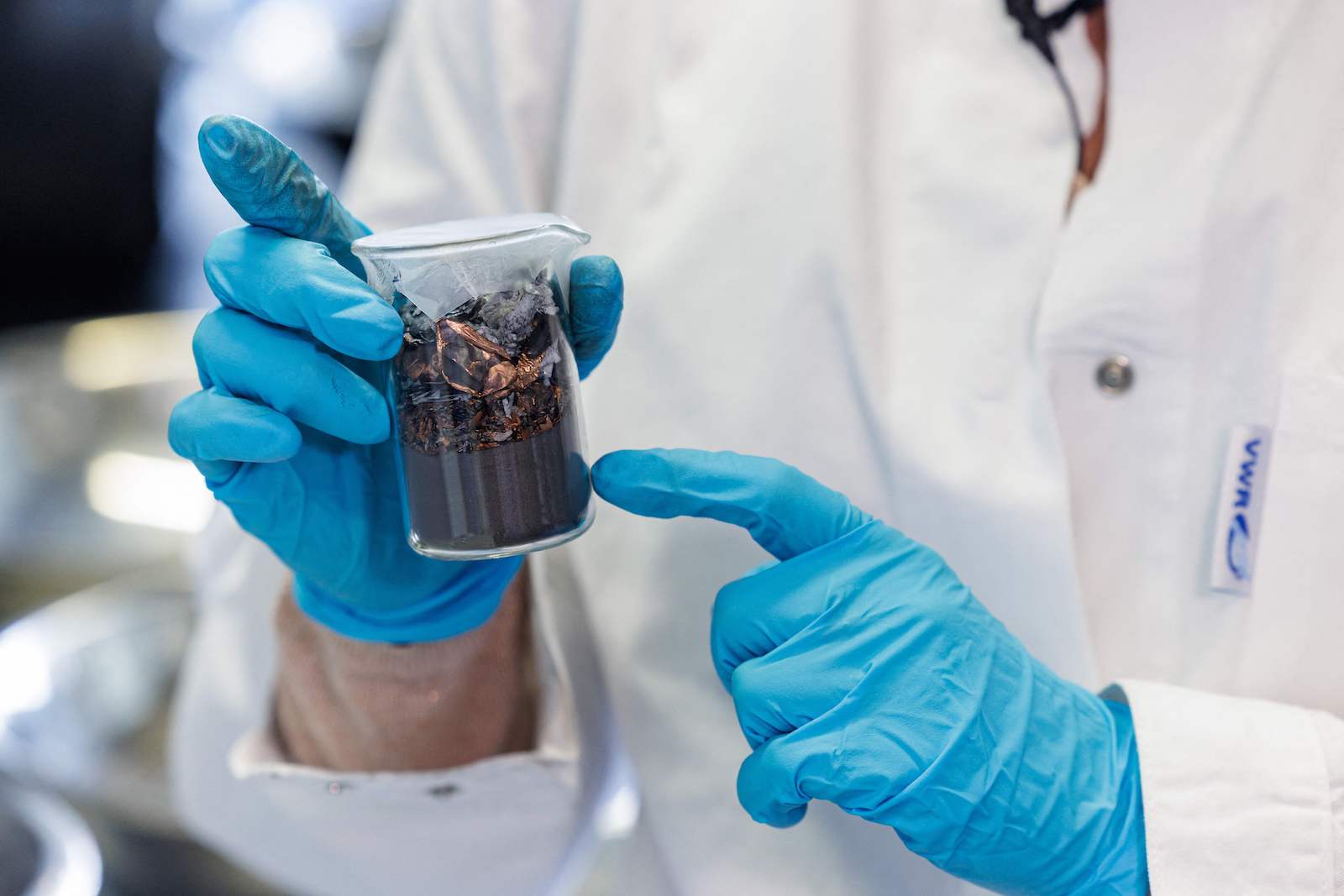 a person with blue gloves holds a beaker filled with sediment of various sizes