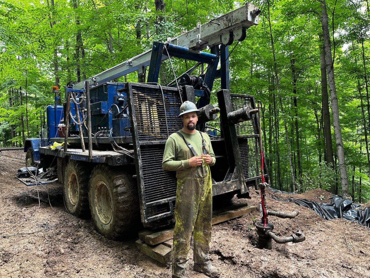 A man in overalls stands next to a large truck in the woods