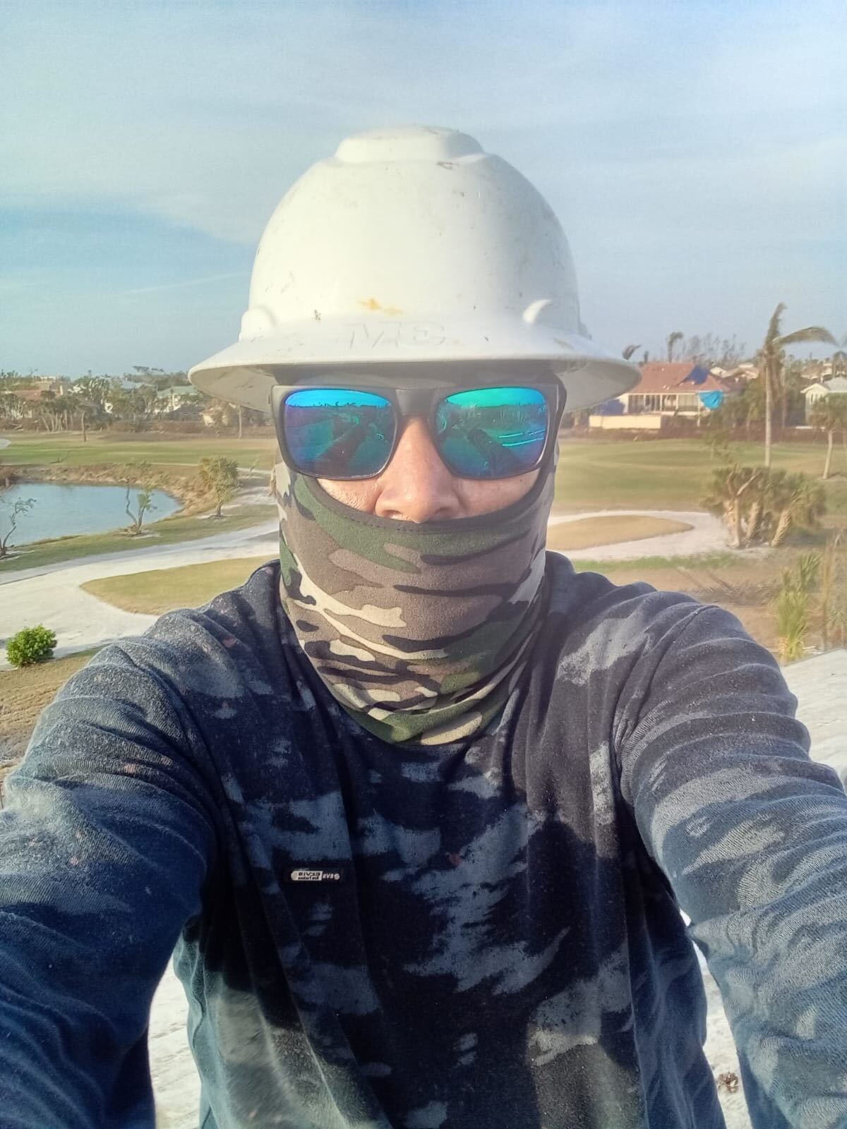 Marcos, a disaster restoration worker in Florida, poses for a selfie at work. He is wearing a white hardhat, sunglasses and a camouflage-print face covering and matching camouflage long-sleeve shirt.