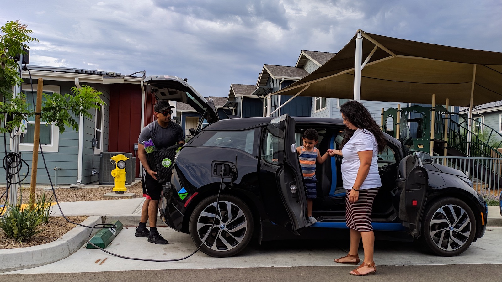 A family of three three is shown unloading groceries in a small BMW electric car plugged in outside their home.